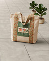 A canvas tote bag with a green front pocket, floral embroidered patch, and patterned side straps, placed on a light gray tiled floor next to two potted fiddle-leaf fig plants.