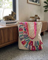 A cream canvas tote bag with vibrant multicolor patchwork embroidery and pink patterned handles, resting on a polished wooden floor next to a wooden sideboard in a modern home interior.